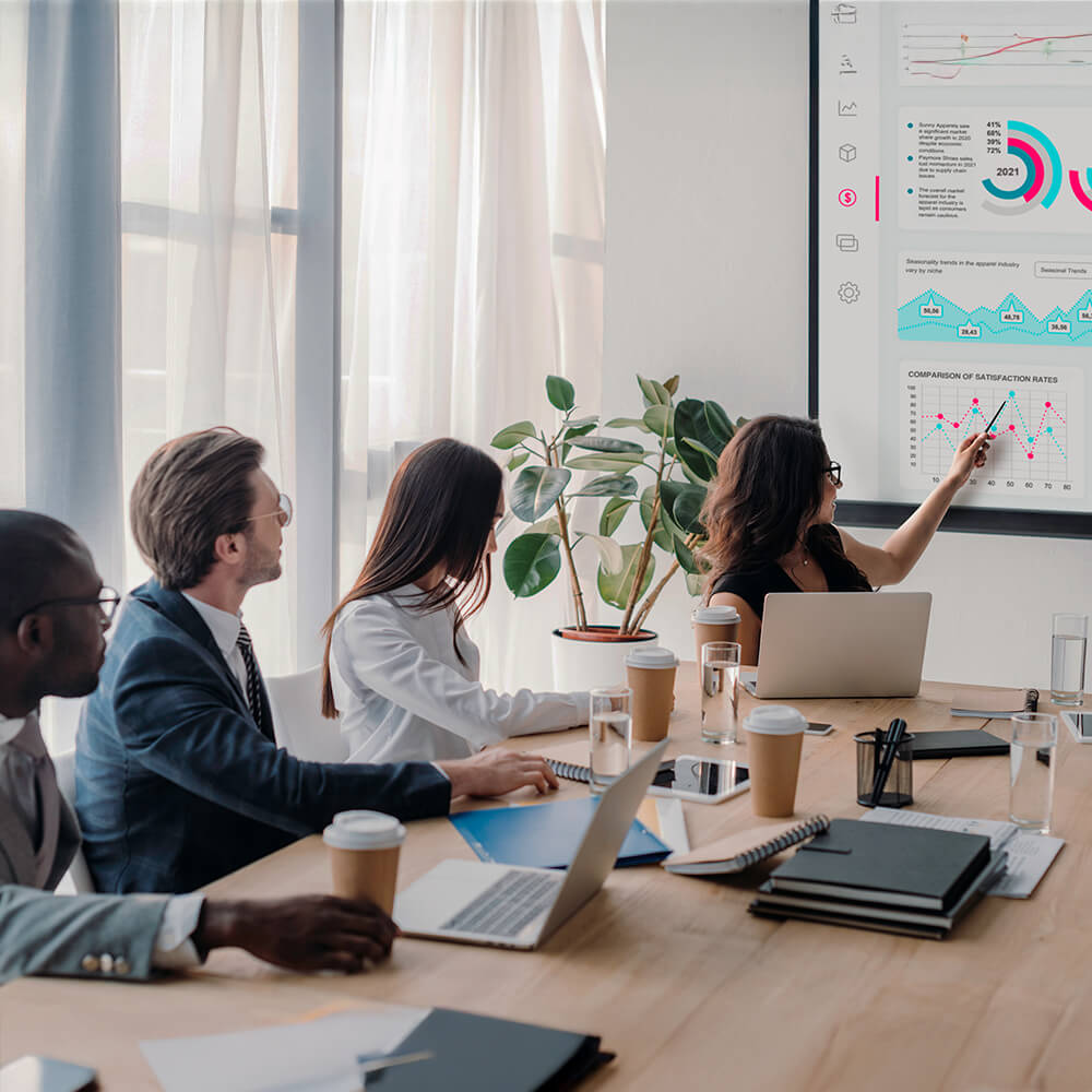 A modern meeting room with people having a meeting sitting around a table. They have laptops and are looking at a large flat panel screen on the wall what is showcasing stats.
